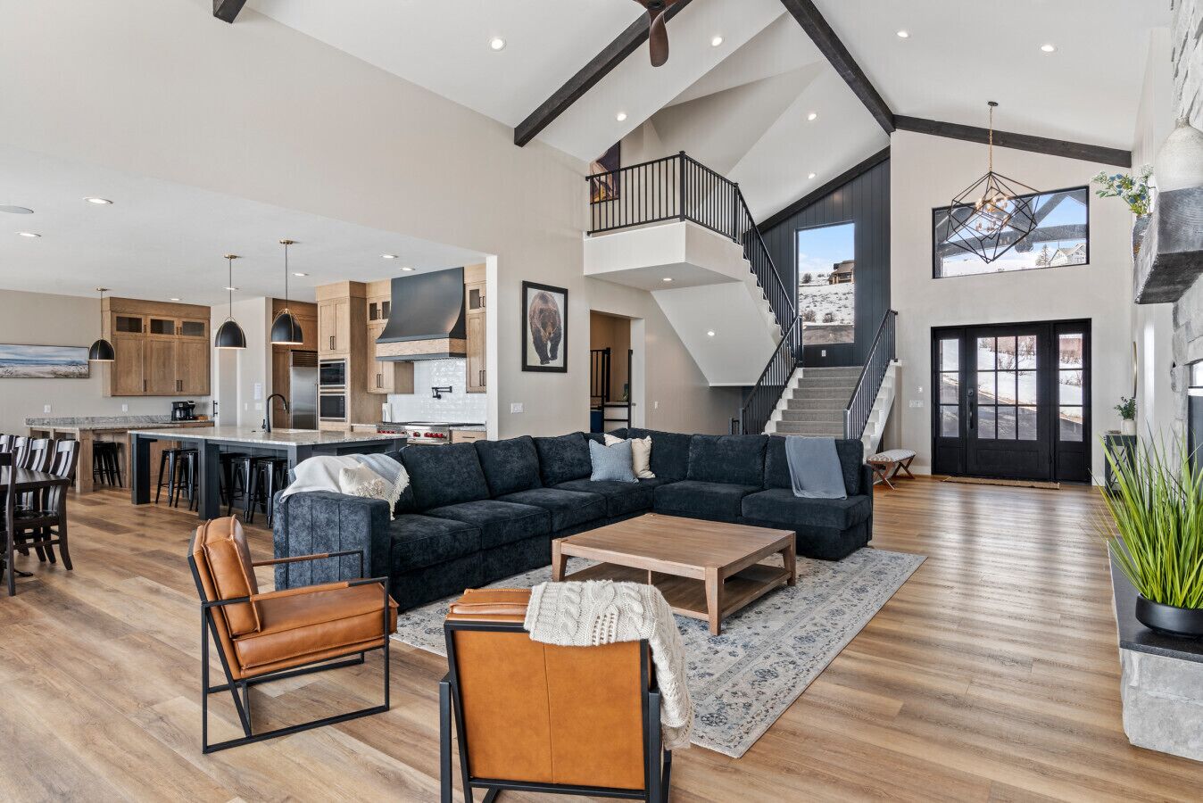 A spacious living area with a large dark-colored sectional sofa, a wooden coffee table, and leather armchairs. A staircase leads to an upper level, and a kitchen with wooden cabinets and a central island is visible.