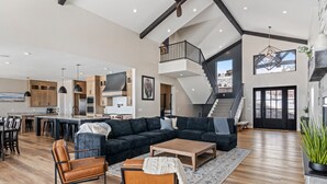A spacious living area with a large dark-colored sectional sofa, a wooden coffee table, and leather armchairs. A staircase leads to an upper level, and a kitchen with wooden cabinets and a central island is visible.