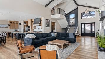 A spacious living area with a large dark-colored sectional sofa, a wooden coffee table, and leather armchairs. A staircase leads to an upper level, and a kitchen with wooden cabinets and a central island is visible.