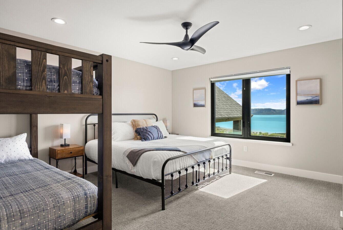A bedroom with a bunk bed, a single bed, a ceiling fan, a window with a view, and a nightstand with a lamp.