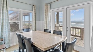 Een eetgedeelte met een grote tafel en stoelen, grote ramen met uitzicht op de oceaan, en een houten terras buiten.