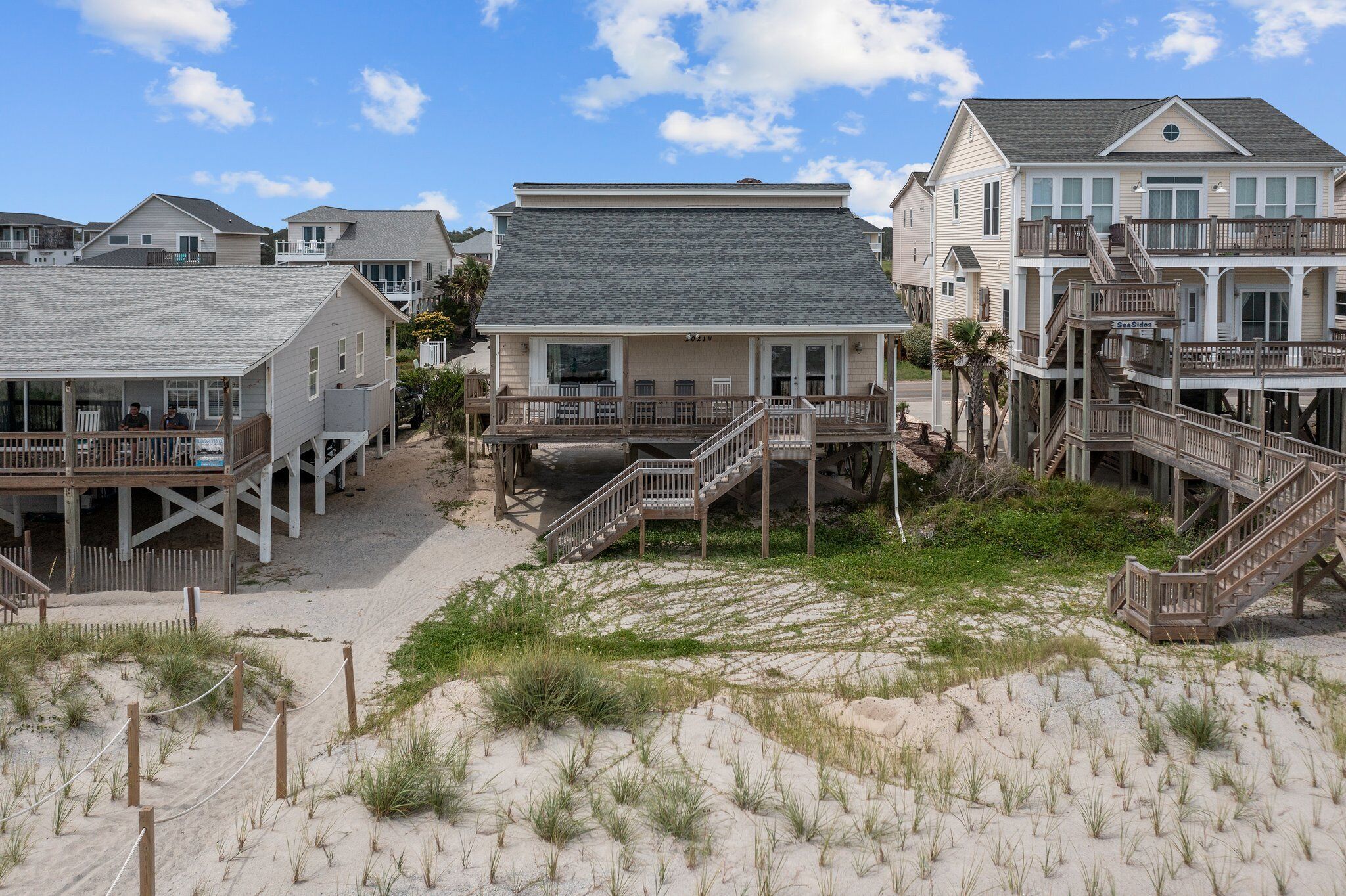 A beachfront property with multiple houses connected by walkways, featuring sandy grounds and grassy areas.