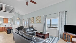Een ruime woonkamer met een grijze modulaire bank, een houten salontafel en een televisie. Er is een balkon met uitzicht op de oceaan, en een keuken is zichtbaar op de achtergrond.