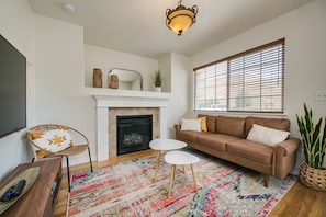 A living room with a fireplace, a brown sofa, a white coffee table, a wicker chair, and a colorful rug. - #HabitueHomes  - The Touchdown - Near BSU! (Boise)