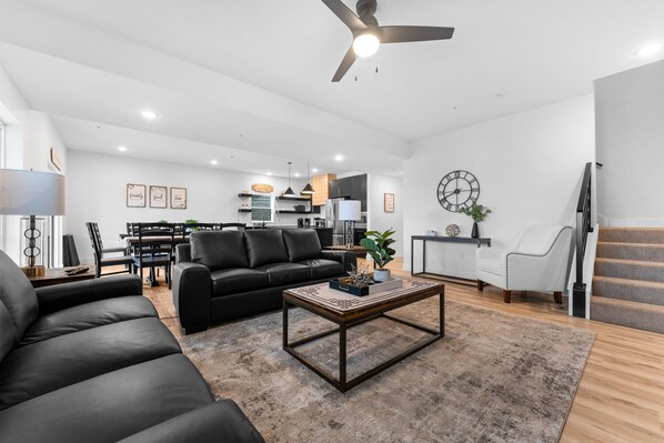 A modern living room with a black leather sofa, a wooden coffee table, and a dining area in the background. - Cedar Shores 7 Bedroom Home by RedAwning (Hollister)