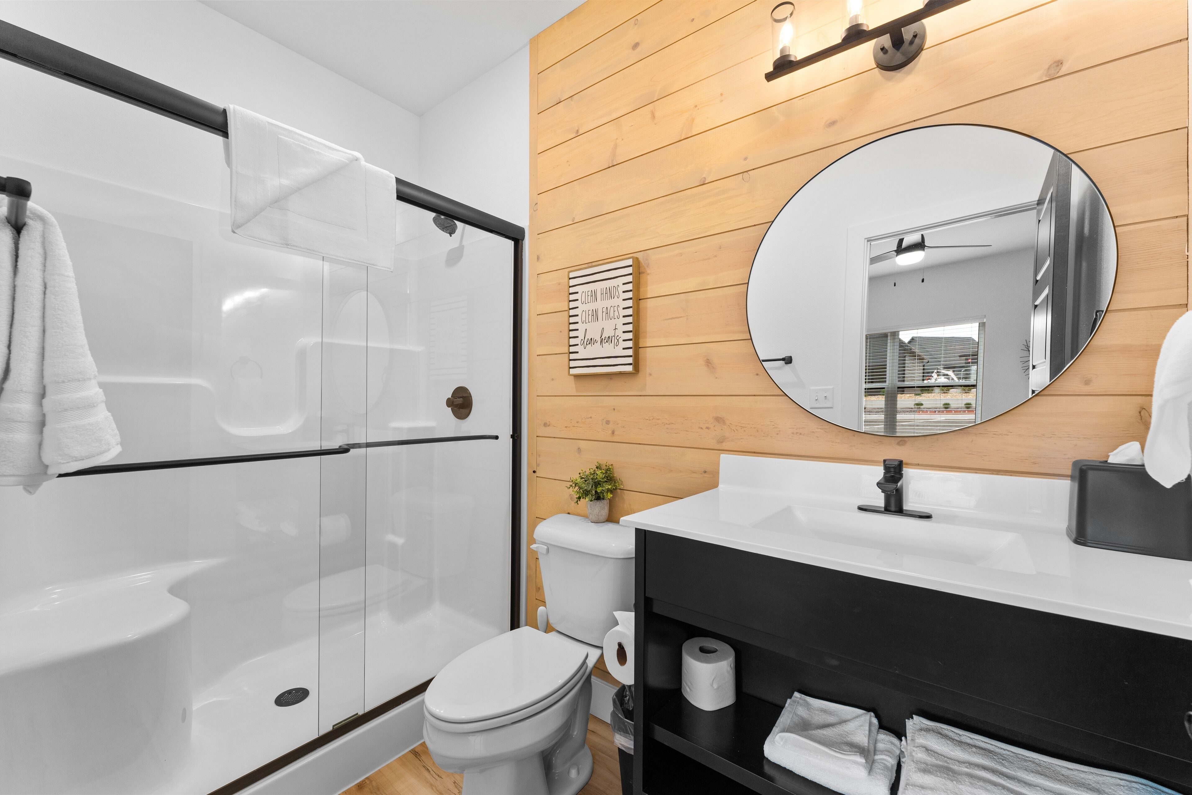 Un baño moderno con cabina de ducha de vidrio, inodoro blanco, tocador negro con espejo y panel de pared de madera.