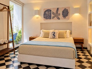 Een hotelkamer met een bed, een houten kapstok, een blauwe stoel, een nachtkastje en een wand met kunstwerk.