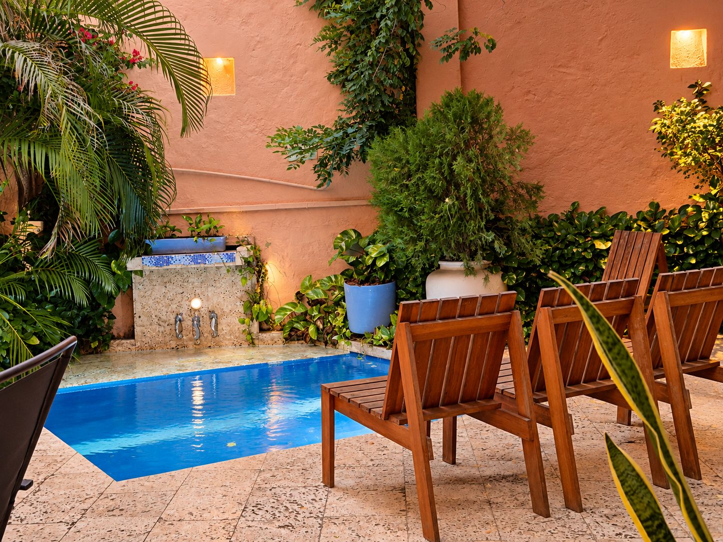 A pool area with a blue pool, wooden chairs, and various potted plants.