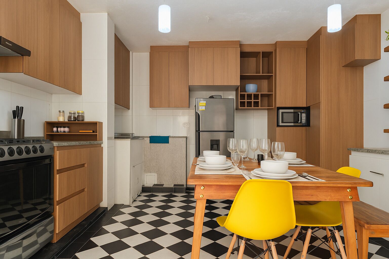 A modern kitchen with wooden cabinets, a black and white checkered floor, a dining table set with white plates and yellow chairs, and a built-in microwave.