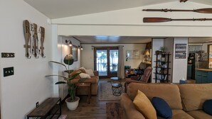 A cozy living room with a brown sofa, a coffee table, and a potted plant. There is a bookshelf with books and a lamp. A wooden sign with the word "RELAX" hangs on the wall.