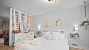 A bedroom with a large bed, white walls, a wooden floor, a white dresser, a mirror, and a hanging plant. - Luxury pet-friendly remodeled cottage (Sparta)