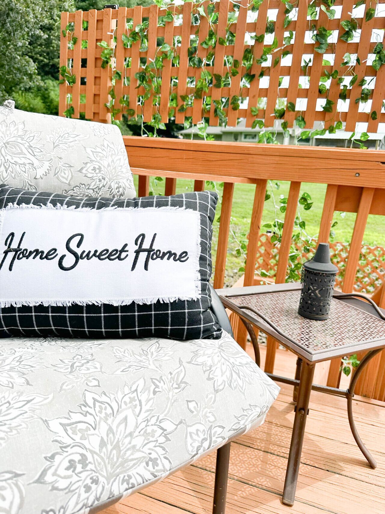 Una terraza de madera con una mesa y sillas de metal, un cojín blanco con el texto 'Home Sweet Home' y una valla de madera con celosías y follaje.