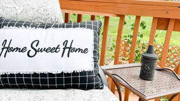 Una terraza de madera con una mesa y sillas de metal, un cojín blanco con el texto 'Home Sweet Home' y una valla de madera con celosías y follaje.