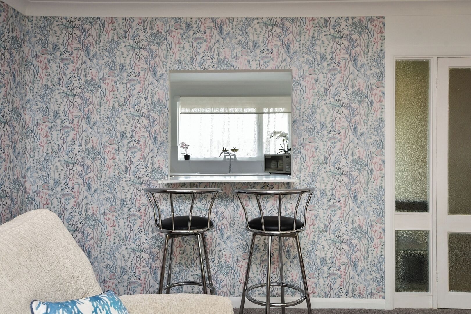 Une pièce avec un papier peint à motifs, un évier blanc, deux tabourets de bar, une chaise blanche et un grand miroir au-dessus de l’évier.