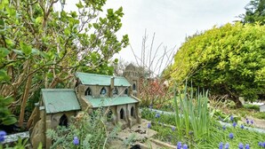 A miniature model of a stone building with arched windows and a green roof, surrounded by lush greenery and purple flowers.