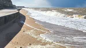 A coastal walkway with a concrete barrier, waves crashing onto the shore, and a cliff in the background.