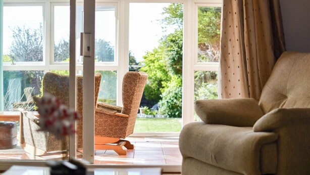 A conservatory with a sofa and armchair, a table with a vase, and a view of a garden.