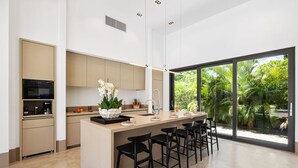 Een moderne keuken met een centraal eiland, zwarte barkrukken en een uitzicht op weelderige begroeiing door grote ramen.