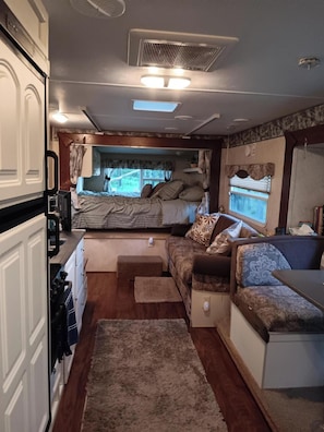 Interior compacto de una caravana con kitchenette, cama, sofá y ventana con vista a zonas verdes.