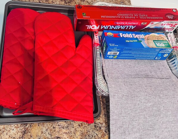 Una encimera de cocina con un guante rojo para el horno, papel de aluminio, una caja de bolsas para sándwiches y una caja azul de bolsas para guardar alimentos.