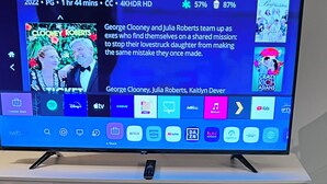 Een televisie op een witte standaard, met een afstandsbediening erboven. Daaronder een glazen tafel met geometrische metalen versieringen en een bordje met de tekst 'Smile'.