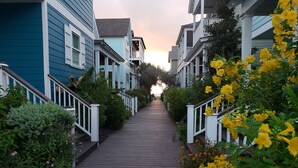 Un chemin de bois mène à une rangée de maisons de ville dotées de balcons blancs, et un coucher de soleil se profile à l’arrière-plan.