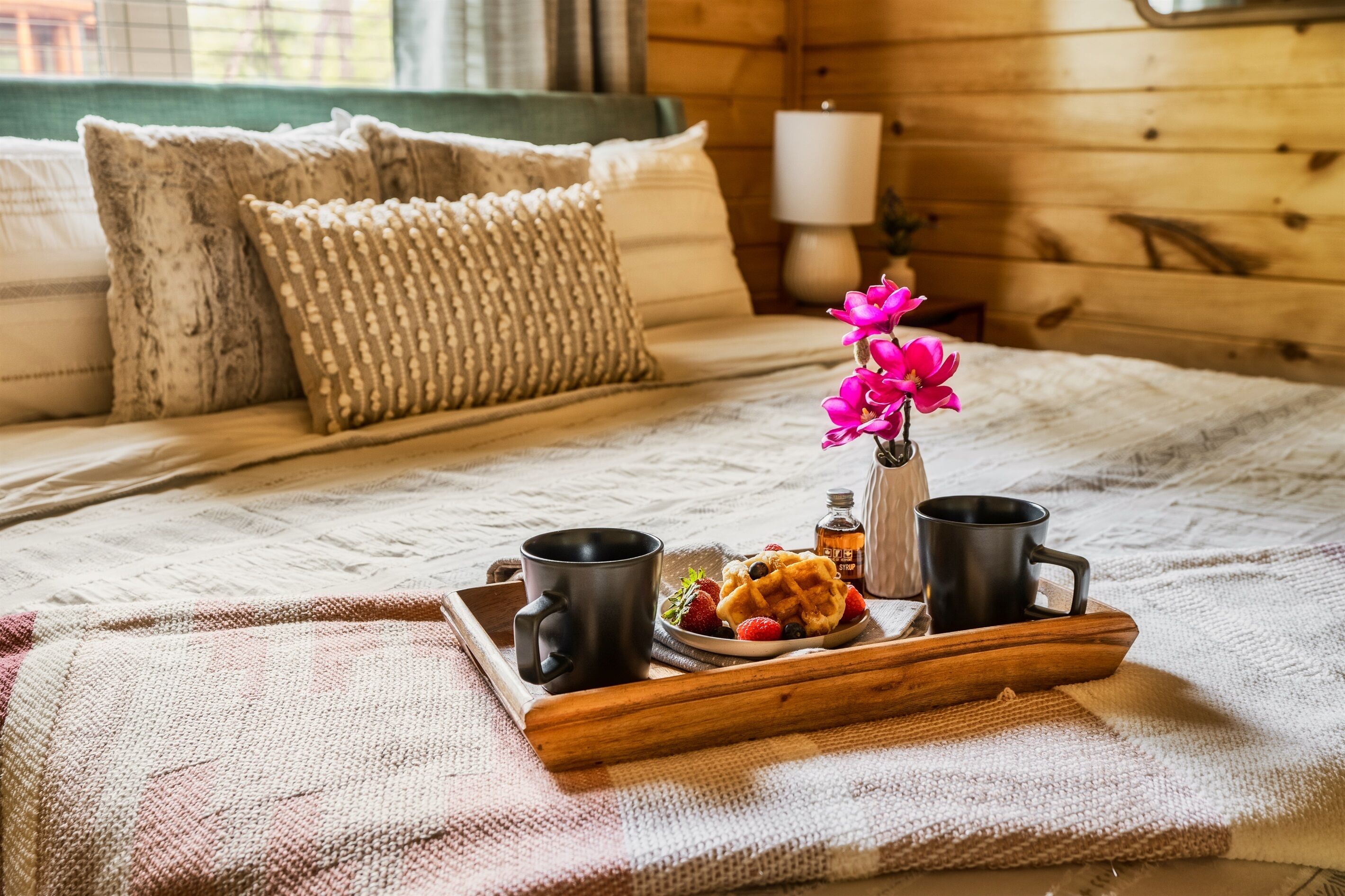 Una cama de madera con colcha de tonos blanco y beige, dos tazas negras, un plato de gofres, fresas y arándanos, y un florero con flores rosadas sobre una bandeja de madera.
