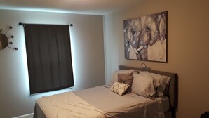 Et soveværelse med en seng, mørke gardiner, et maleri og en vægmonteret lampe.