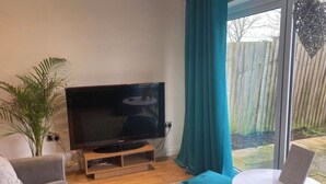 A living room with a flat-screen TV, a grey sofa, a small round table with a remote, and a large window with curtains. - Lovely 5-Bed House in Brighton (Brighton)