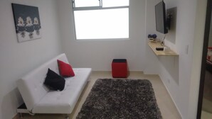 A modern living room with a white sofa, red and black cushions, a dark rug, a small red ottoman, a flat-screen TV on a wooden shelf, and a window with natural light. - Great Location: Apartment Nearby Itagui Metro Station (Sabaneta)