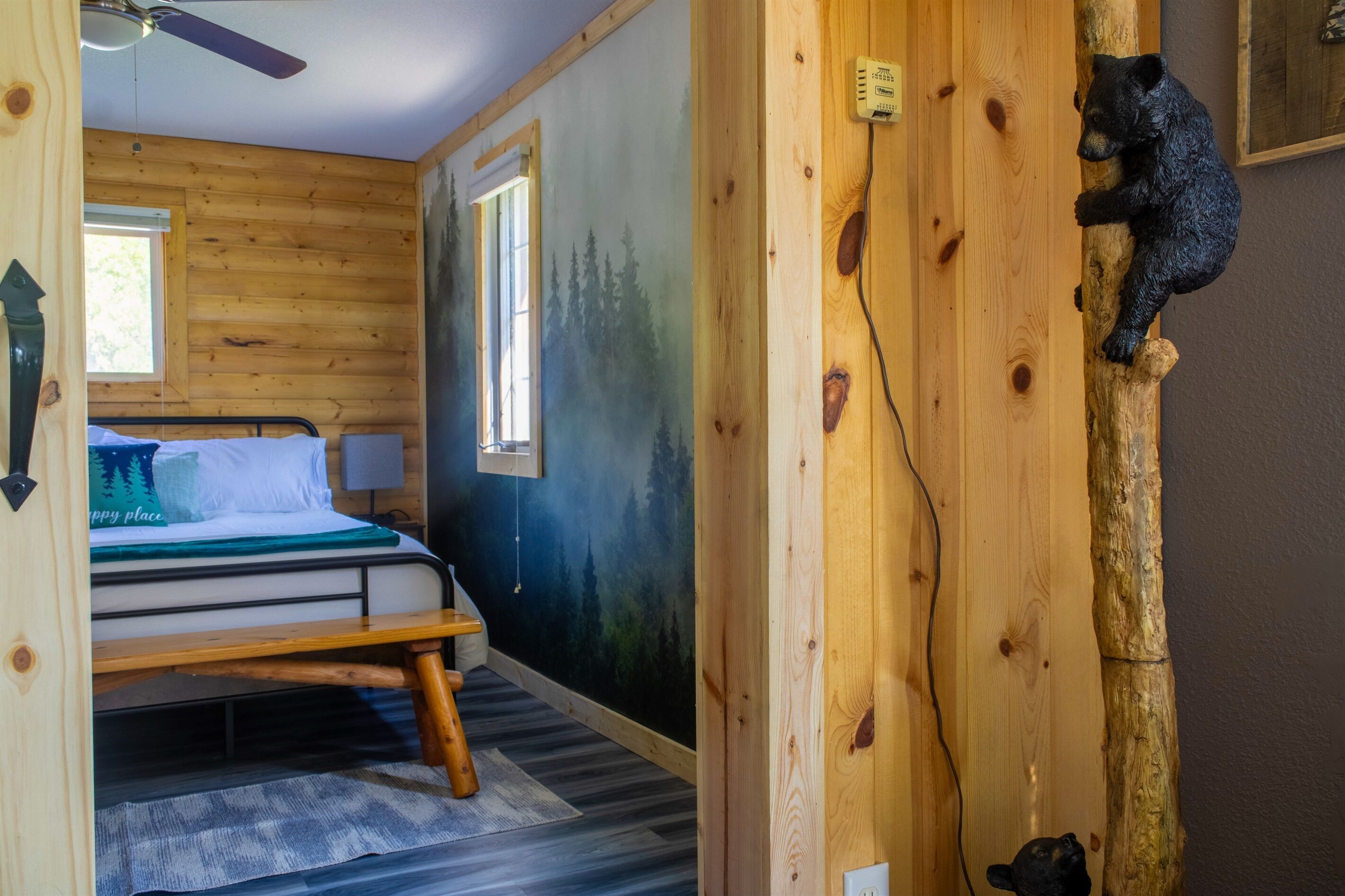 A wooden cabin room with a bed, a window, a wall-mounted light, and a bear sculpture.