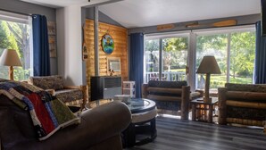 Ein gemütliches Wohnzimmer mit Kamin, bequemen Sitzmöglichkeiten, einem Couchtisch und großen Fenstern mit Blick ins Freie.