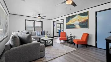 Un salon moderne avec un canapé gris, une table basse et deux fauteuils orange. Un tableau est accroché au mur et un ventilateur de plafond est installé.
