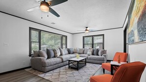 Un salon moderne avec un canapé d’angle, une table basse et un fauteuil orange vif. Il y a deux ventilateurs de plafond et de grandes fenêtres équipées de stores.