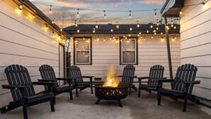 Une terrasse avec un foyer et des fauteuils Adirondack, des guirlandes lumineuses suspendues au-dessus, et une maison avec deux fenêtres.