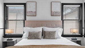 Une chambre à coucher avec un grand lit, deux tables de chevet et deux fenêtres équipées de stores.
