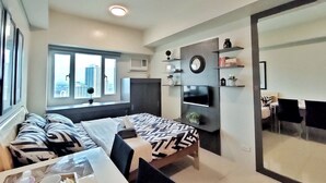 Een moderne hotelkamer met een bed, een eettafel met stoelen, een aan de muur bevestigde televisie en een groot raam met uitzicht over de stad.