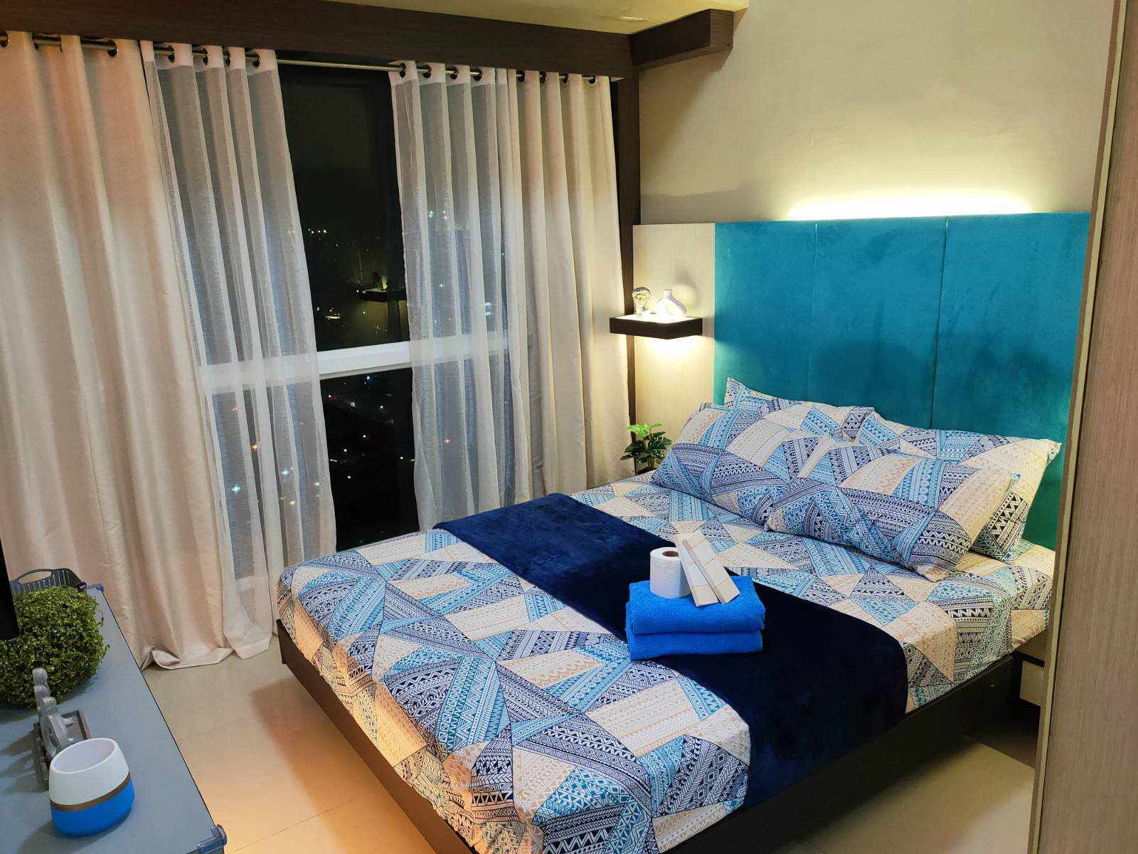 A neatly made bed with patterned bedding, a bedside lamp, and a view of the city at night through sheer curtains.