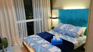 A neatly made bed with patterned bedding, a bedside lamp, and a view of the city at night through sheer curtains.