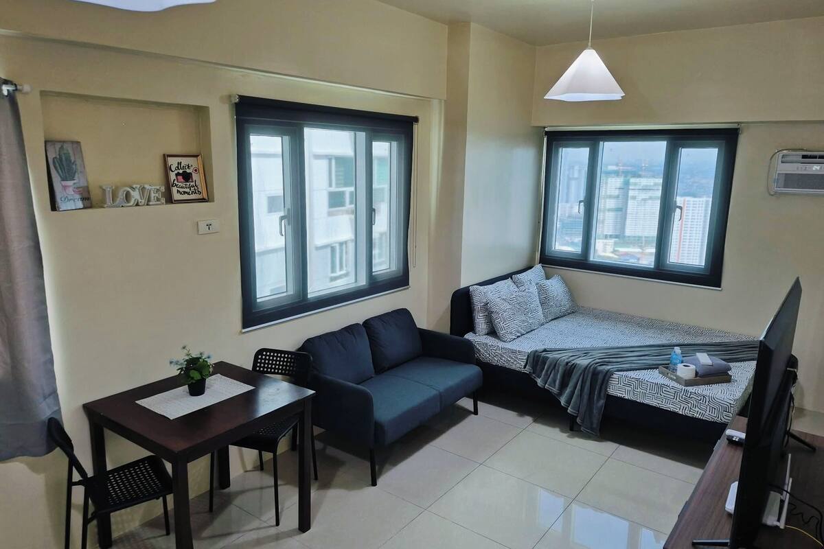Ein gemütlicher Raum mit Bett, Sofa, kleinem Tisch mit Pflanze, Fernseher und großen Fenstern.