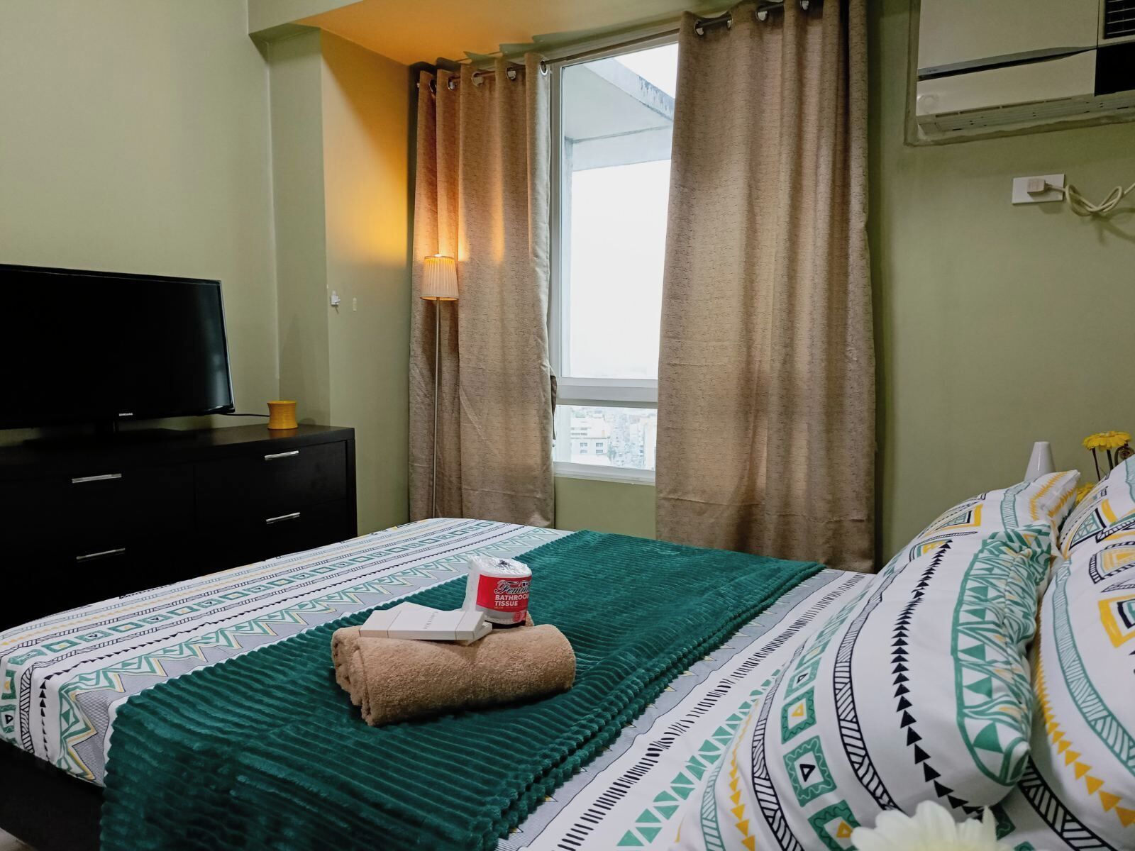 A bedroom with a bed, a TV, a window with curtains, and an air conditioning unit.