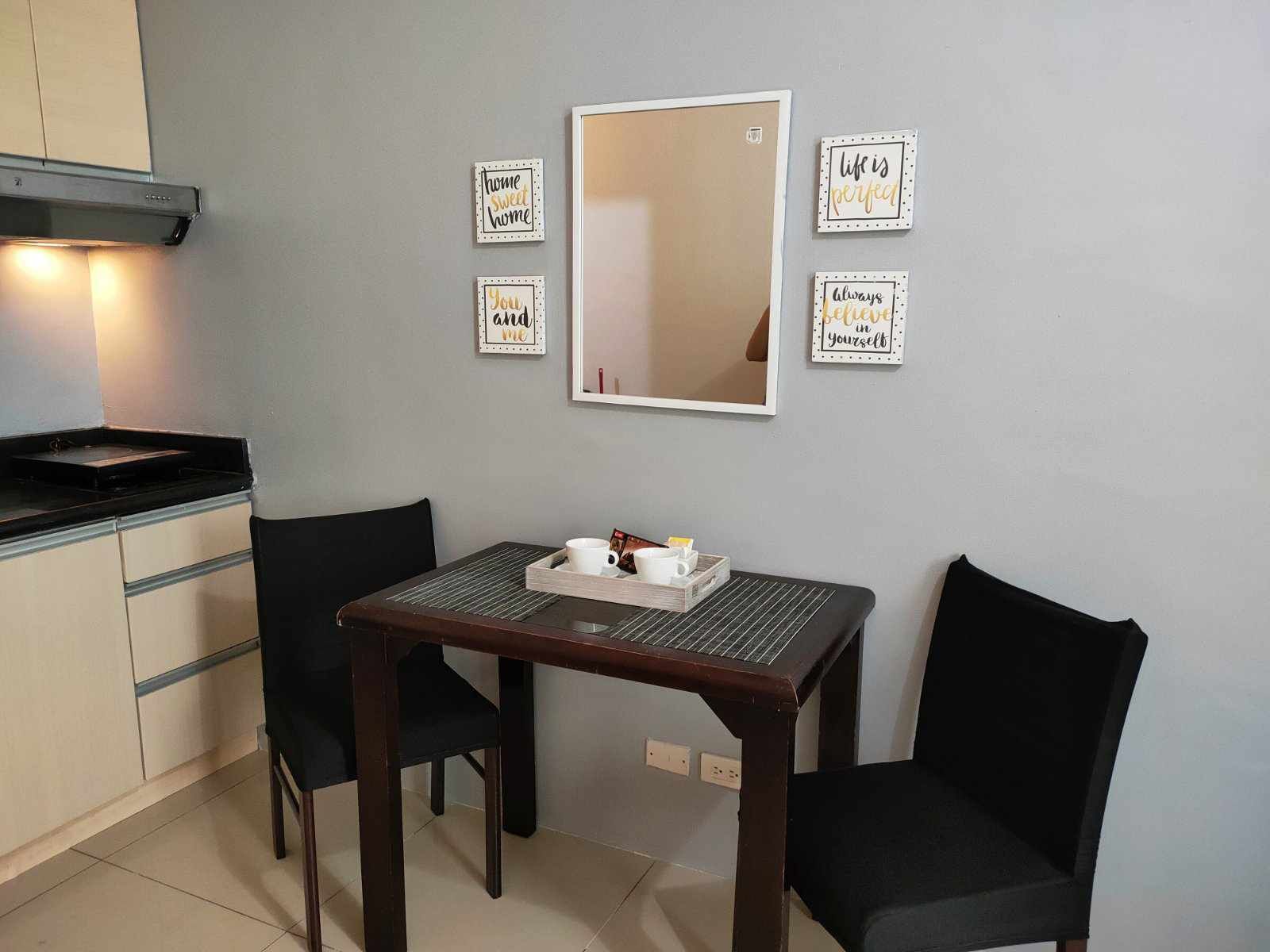 A small kitchenette with a stove, a sink, and a countertop. There is a table with a tray of items, two chairs, and a mirror on the wall.