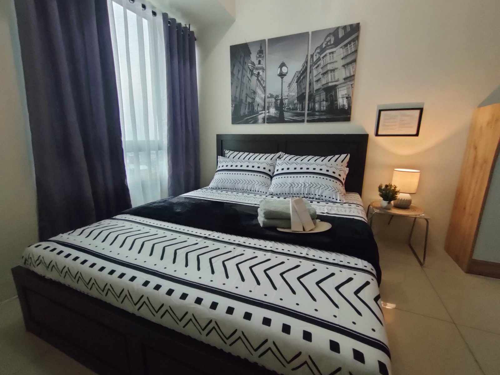 A neatly made bed with patterned bedding, a bedside table with a lamp, and a window with curtains.
