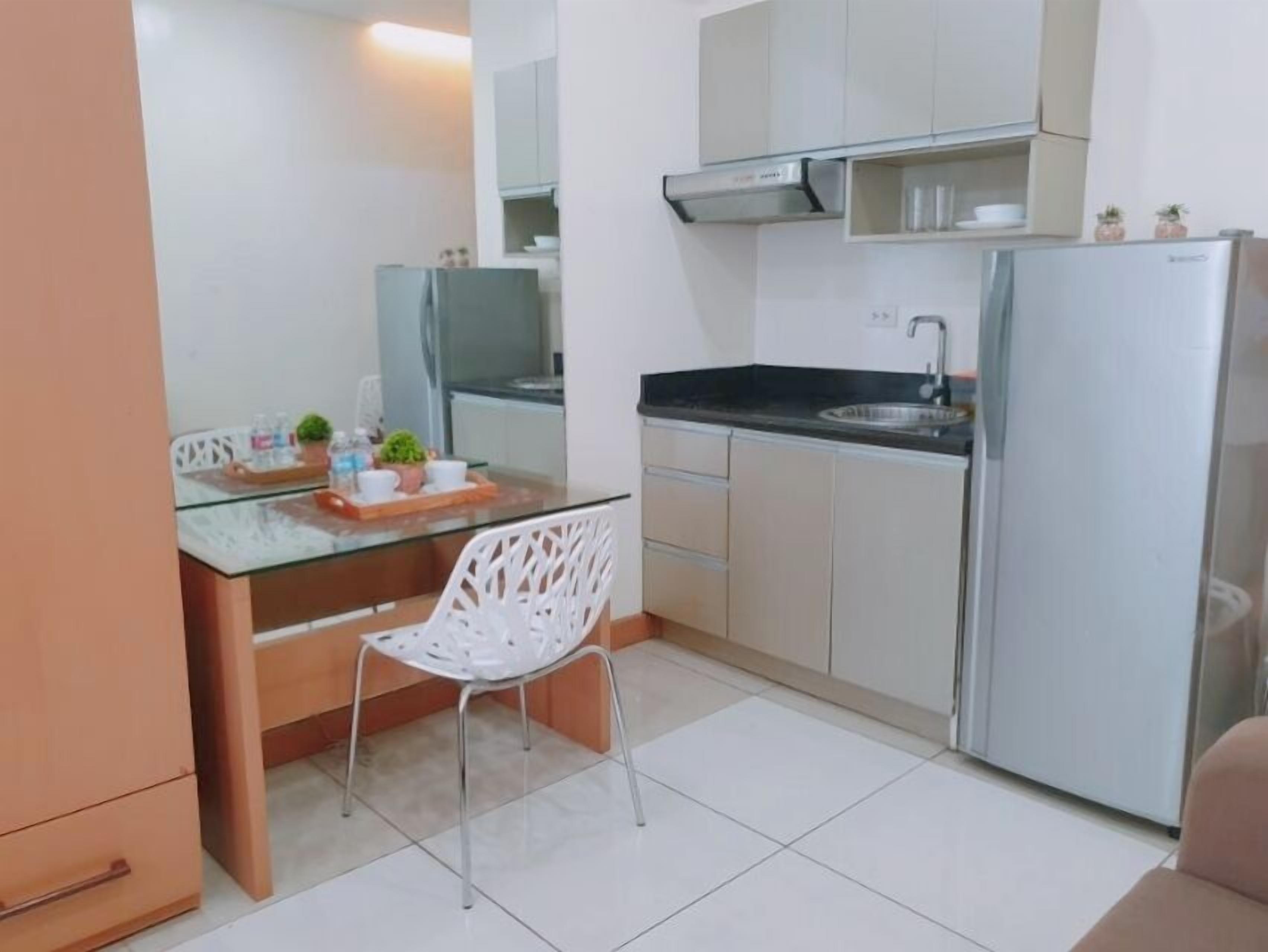 A compact kitchen with a glass-top dining table, white chairs, a built-in oven, and a refrigerator.