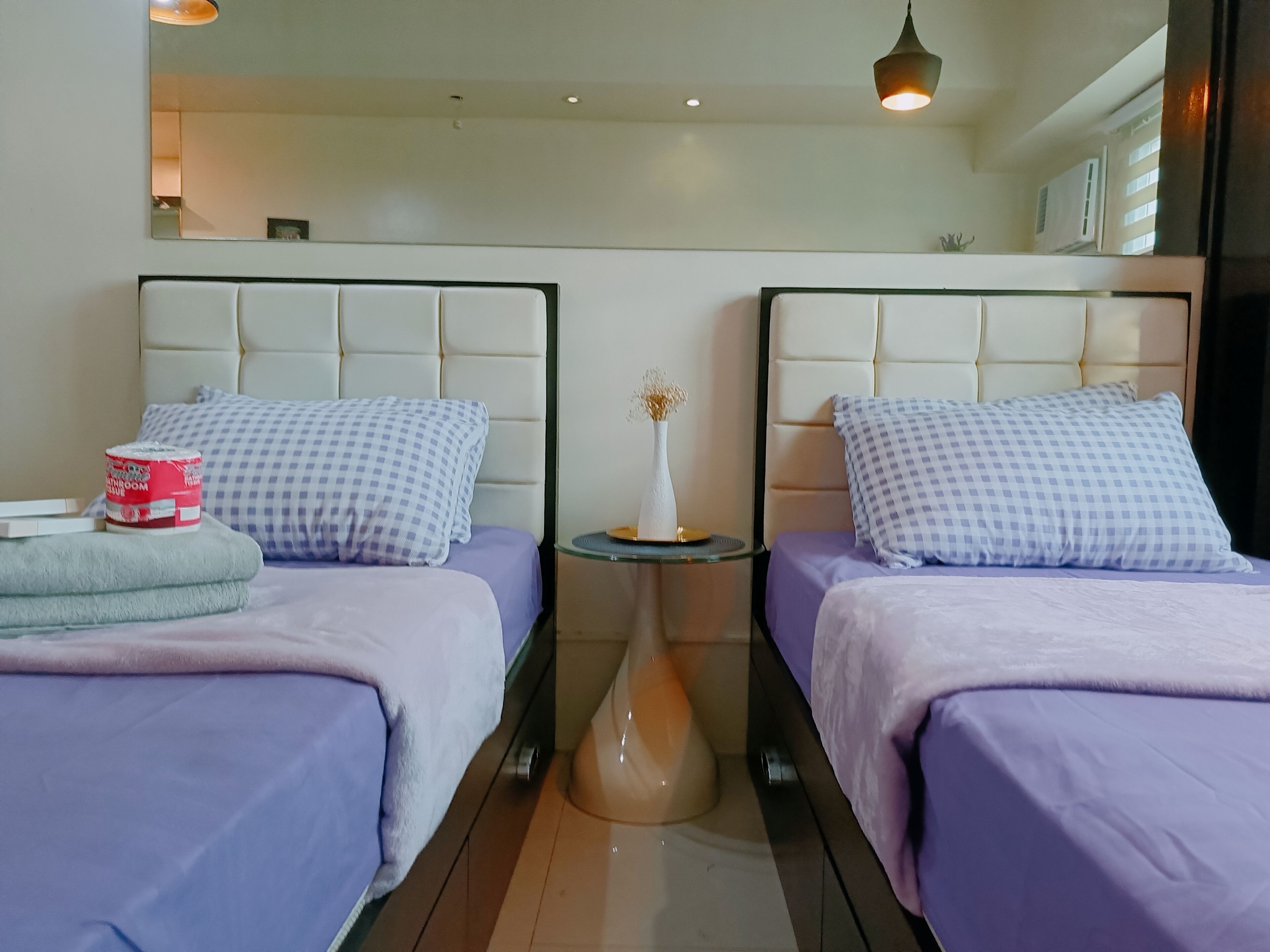 A hotel room with two beds, each with a checkered pillow and a purple bedspread. There is a small table between the beds with a vase and a book.