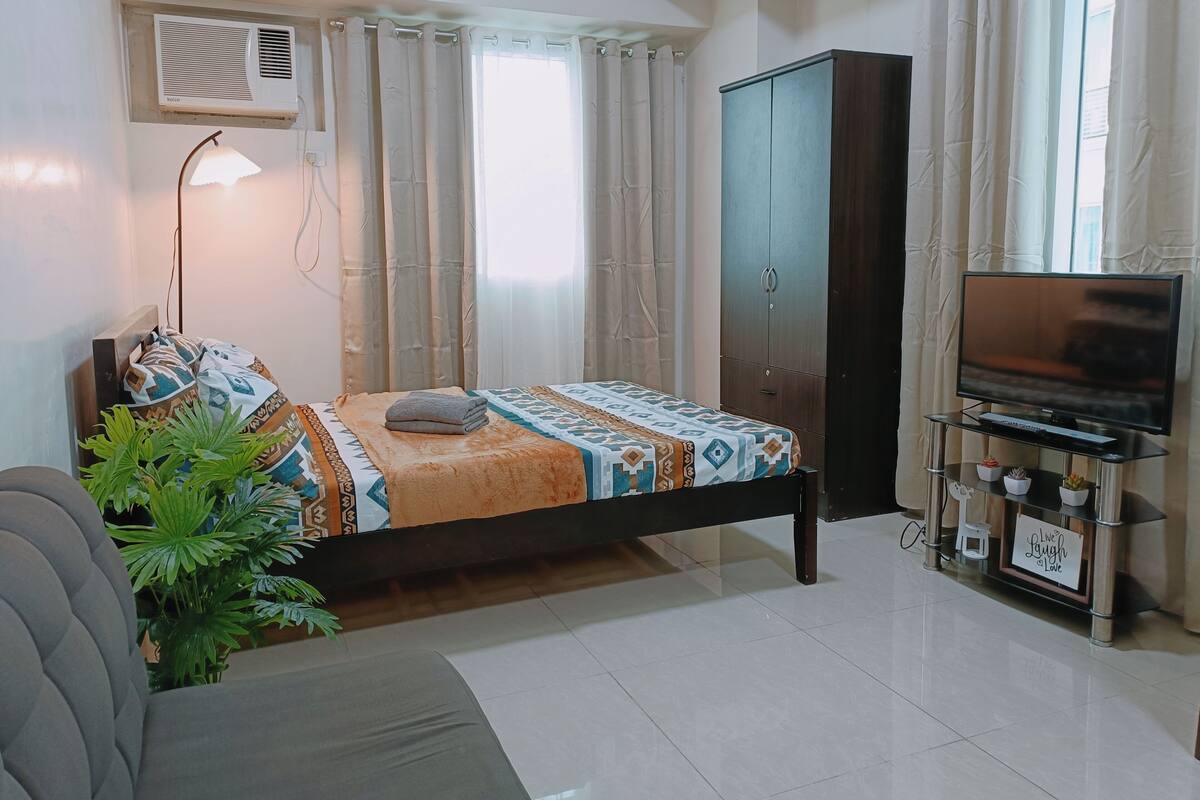 Ein Schlafzimmer mit einem Bett, einer TV-Kommode mit Flachbildfernseher, einem Kleiderschrank, einer Lampe und einer Zimmerpflanze.
