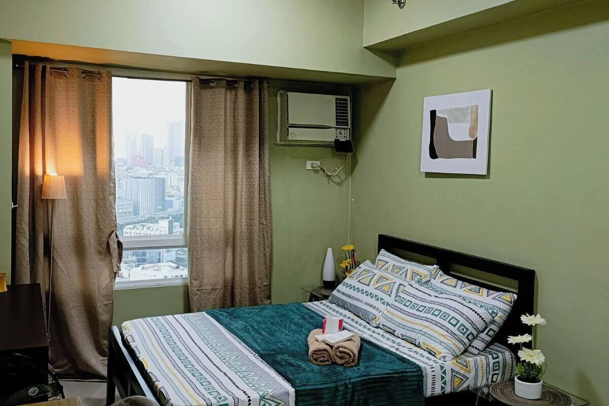 Ein Hotelzimmer mit Bett, Schreibtisch, Stuhl, einem Fenster mit Vorhängen, einer Klimaanlage und Blick auf die Stadt.