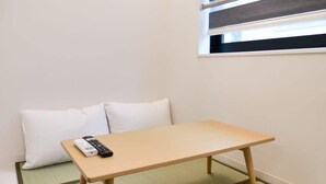 A small room with a wooden table, two white pillows, a remote control, and a window with blinds. - 2023 Open/Shinjuku Okubo/5min 2 JR/Premium Capsule (Shinjuku City)