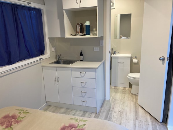 A compact kitchen with a sink, a countertop, and cabinets. There is a small window with a blue curtain, a mirror above the sink, and a door leading to a bathroom with a toilet and sink. - Kiwi Bach with Cabin and room for your boat.
15 min walk to the beach.
(Ruakākā)