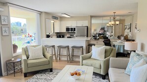 Ein modernes Wohnzimmer mit einer Küche im Hintergrund, ausgestattet mit einem Essbereich mit Tisch und Stühlen, einem Sofa und einem Couchtisch mit einer Obstschale.